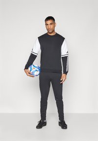 Zwart-wit sport sweatshirt met lange mouwen, gecombineerd met zwarte, nauwsluitende broek. Houdt een kleurrijke, gestructureerde voetbal vast.