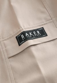 Étiquette en tissu beige avec lettres noires indiquant « BAKER by TED BAKER », comprenant les informations de taille et d'entretien, fixée à une surface texturée.