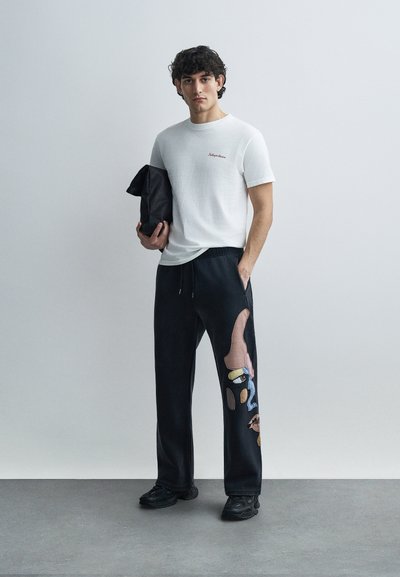 Λευκό μπλουζάκι με υφή και λογότυπο, συνδυασμένο με μαύρη φόρμα με πολύχρωμη αφηρημένη κέντημα και ελαστική μέση. Μαύρα παπούτσια.