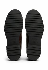 Schwarze Gummisohle mit geriffelter Struktur, mit einem prägenden Tommy Jeans-Logo an der linken Ferse, verbunden mit dem oberen Teil aus braunem Leder.