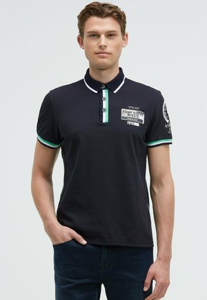 Junger Mann trägt ein marineblaues Polohemd mit grüner und weißer Einfassung, Knopfleiste und Patches auf Brust und Ärmel, steht mit der Hand in der Tasche.