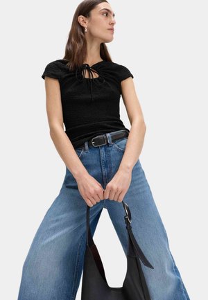 Vrouw draagt een zwart gestructureerd T-shirt met korte mouwen en wijde blauwe jeans, houdt een zwarte leren handtas vast voor een lichtgrijze achtergrond.