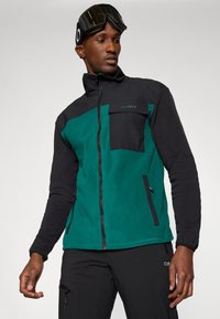 Chaqueta de forro polar en verde y negro, con un alto cuello, cierre de cremallera en la parte delantera, bolsillo en el pecho, bolsillos laterales y texturas contrastantes.