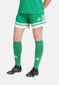 Grüne Fußballshorts mit weißen Streifen, kombiniert mit grünen Kniestrümpfen. Schwarze Fußballschuhe haben ein strukturiertes Design und weiße Akzente.