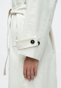 Lysebeige trenchcoat med tekstureret stof, der har en foldbar manchet og to store sorte knapper, og som lukkes med et matchende bælte.