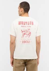 T-shirt en coton beige avec texte et graphiques imprimés en rouge au dos, incluant un cheval et l'année "1932", à manches courtes et col rond.
