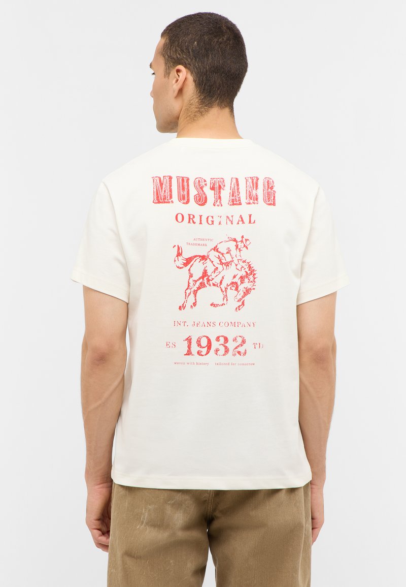 T-shirt en coton beige avec texte et graphiques imprimés en rouge au dos, incluant un cheval et l'année "1932", à manches courtes et col rond.