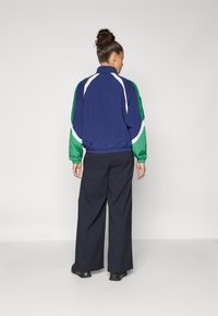 Giacca a vento blu navy con maniche verdi, dettagli bianchi e polsini elasticizzati. Viene indossato un pantalone nero a gamba larga e scarpe nere.