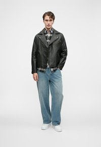 Jaqueta de motociclista preta de couro com bolsos com fecho, usada sobre uma camisa xadrez, combinada com jeans soltos de azul-claro e ténis brancos.