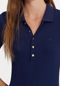 Navyblå pikétröja i ribbat tyg med knäppning i tre knappar, utrustad med guldtonade knappar och en liten logotyp på vänster bröst.