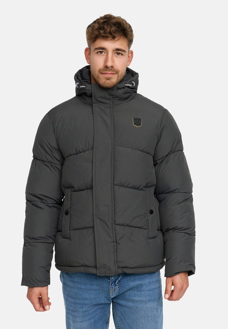 Jonge man met lichtbruin haar, gekleed in een zwarte gewatteerde jas met capuchon en blauwe jeans, staand tegen een effen lichte achtergrond.