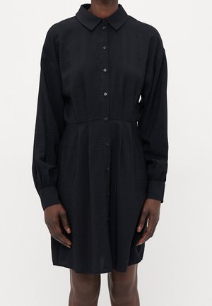 Zwarte, lange mouwen, overhemdjurk met knopen en een kraag, gedragen door een persoon die met ontspannen armen langs het lichaam staat.