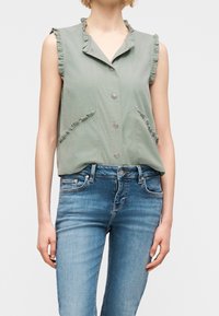 Haut vert sans manches à boutons avec un volant au niveau du col et des poches, porté avec un jean ajusté bleu clair.