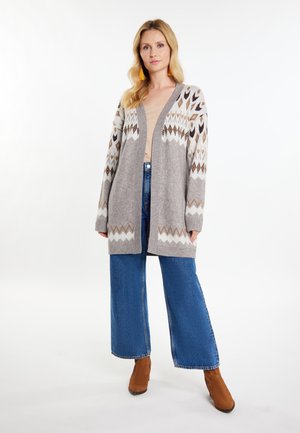 Grauer Strickcardigan mit geometrischen Mustern in Weiß und Braun, offene Vorderseite, getragen über einem beigen Oberteil, kombiniert mit weit geschnittenen blauen Jeans.