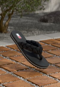 Svart flip-flop med flätad rem. Tjock sula, texturerad botten och logotyp i röd, vit och blå. Står mot en mönstrad stengrund.