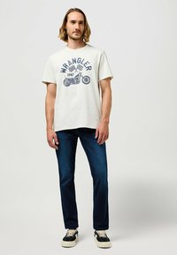 Camiseta de algodón blanca con el gráfico azul "WRANGLER 1947", que presenta una motocicleta y banderas a cuadros. Combinada con jeans azul oscuro y zapatillas.