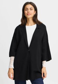 Black wool-blend oversized blazer with a single button closure, notch lapels, and wide sleeves, layered over a white top with lace trim.