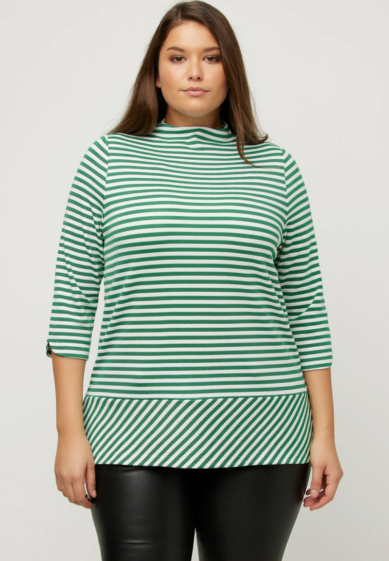 Grün-weißer gestreifter Rollkragenpullover mit Dreiviertelärmeln, glatter Textur und schrägen Streifen am Saum. Kombiniert mit schwarzen Hosen.