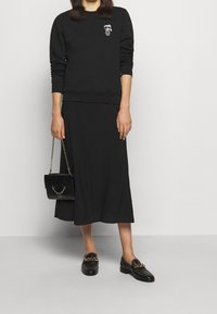 Svart sweatshirt med ett tygmärke, kombinerad med en svart midikjol. Skon är svarta loafers med en guldkedja som detalj. Svart handväska med kedjehandtag.