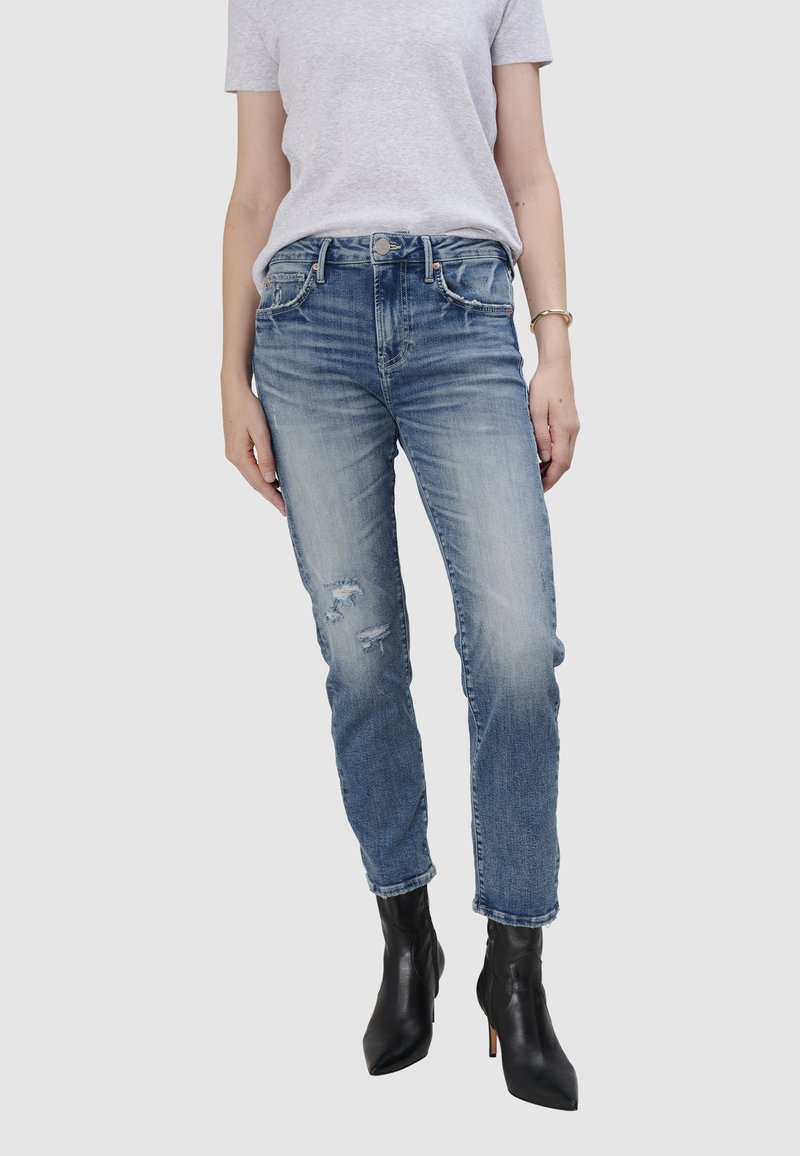 Niebieskie dżinsy o tapered fit, z przetarciami i wyblakłym wykończeniem, zestawione z czarnymi botkami za kostkę i szarym t-shirtem.