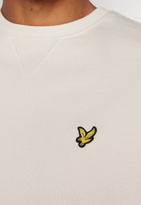 Sudadera blanca con cuello redondo acanalado y pequeño logotipo de pájaro amarillo bordado en el pecho.