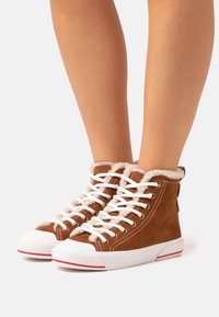 Brązowe zamszowe wysokie trampki z białymi sznurowadłami, futrzanym wykończeniem i białymi gumowymi podeszwami z czerwonymi akcentami. Widoczne złote oczka.