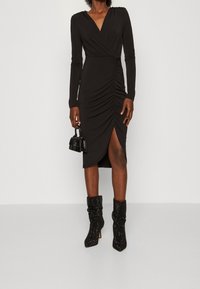 Czarna sukienka z długim rękawem, zdobiona marszczeniami, z dekoltem w kształcie V oraz bocznym rozcięciem. Stylizowana z małą torebką i czarnymi, błyszczącymi botkami za kostkę.