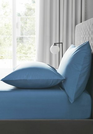 Bett mit blauen Laken und Kissen neben einem grauen gepolsterten Kopfteil und einer runden weißen Nachttischlampe an einem Fenster mit weißen Vorhängen.