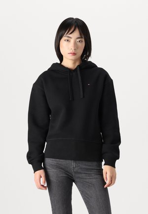 Svart hettegenser med ribbestrikkede mansjetter og nederdel. Har en liten logo på brystet. Kombinert med mørkegrå jeans. Mykt materiale.