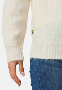 Krämfärgad stickad tröja med ribbade ärmslut och nederkant, med en liten svensk flagga som detalj. Matchad med blå denimjeans.
