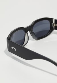 Black sunglasses with a thick, textured frame and rounded lenses. Features gray tinted lenses and subtle branding on the side.