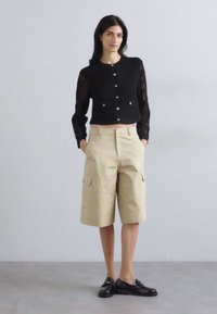 Cardigan noir en tricot avec des manches longues et une fermeture à boutons, associé à un short cargo beige avec des poches avant, et des chaussures noires à enfiler.