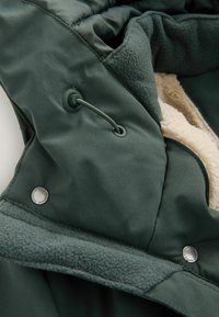 Jachetă pufoasă de culoare verde închis, cu căptușeală din fleece. Dispune de un sistem de închidere cu șnur și nasturi metalici argintii. Interior moale și texturat.