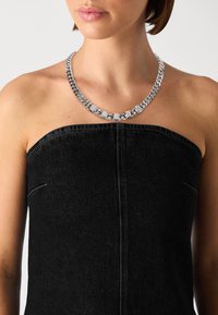 Metal chain necklace with silver finish, featuring block letters spelling "GUES," against a black strapless top. Textured links visible.