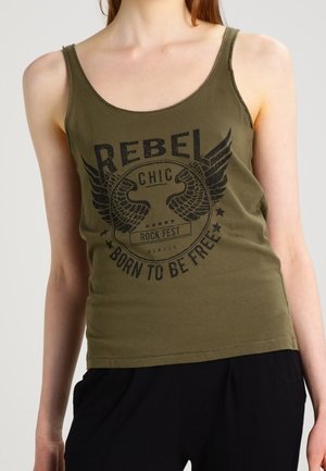 Kobieta nosząca oliwkowo-zielony top z czarnym skrzydlatym emblematem i tekstem "REBEL CHIC ROCK FEST BORN TO BE FREE."