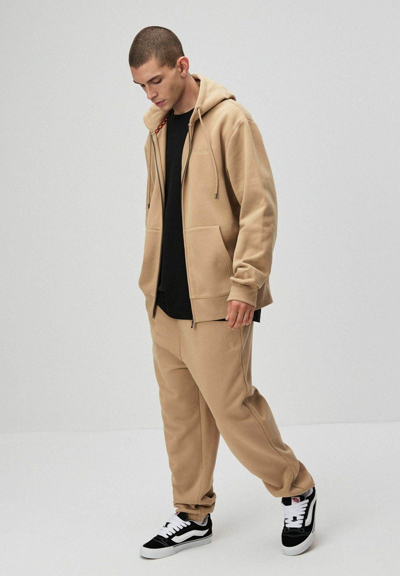 Beżowa bluza z kapturem, z zamkiem i kieszeniami z przodu, zestawiona z pasującymi spodniami dresowymi. Czarna koszulka pod spodem, czarno-białe sneakersy.
