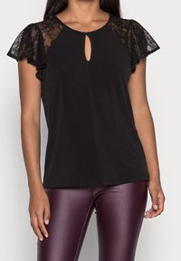 Woman wearing a black top with lace flutter sleeves and a keyhole neckline, paired with shiny maroon leggings, against a neutral background.