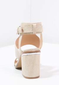 Vaaleanbeige korkokenkä, jossa on teksturoitu päällinen, säädettävä nilkkahihna ja lohkokorko, jossa on hopeinen korostus reunoilla.