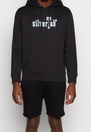 Homme portant un sweat à capuche noir avec le logo « silverTab » en blanc sur la poitrine et un short noir, debout devant un fond clair uni.