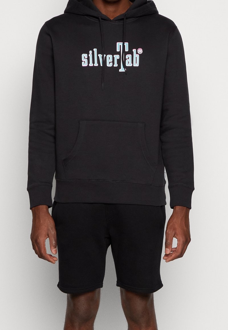 Homme portant un sweat à capuche noir avec le logo « silverTab » en blanc sur la poitrine et un short noir, debout devant un fond clair uni.