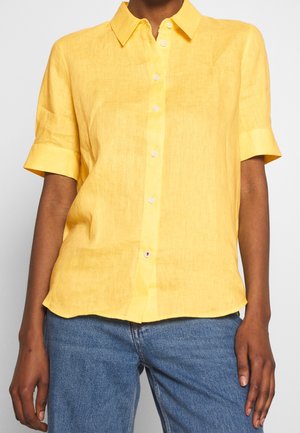 Personne portant une chemise jaune à manches courtes boutonnée associée à un jean en denim bleu, montrée des épaules jusqu'aux hanches.