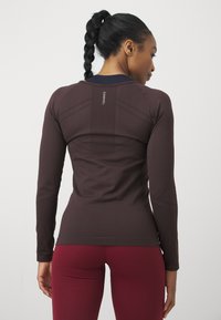 Brūns garas piedurknes sporta tops ar pieguļošu dizainu. Iezīmēts ar teksturētu muguras paneļa un plakaniem šuvēm komfortam. Kombinēts ar sarkaniem legingiem.