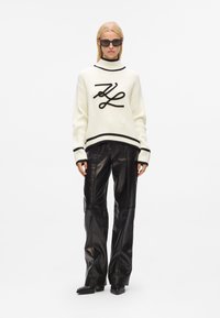 Pull en maille blanche avec des accents noirs, présentant un design de logo central. Associé à un pantalon en cuir noir ample et des lunettes de soleil carrées.
