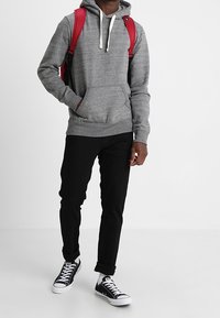 Sudadera gris con un bolsillo en la parte frontal y capucha con cordón, combinada con jeans negros y zapatillas de lona bajas en blanco y negro. Mochila roja sobre los hombros.