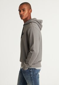 Grijze hoodie met een zwart logo-embleem, voorzien van een verstelbare capuchon, lange mouwen en een relaxed fit. Gecombineerd met blauwe jeans.