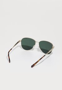 Lunettes de soleil avec monture métallique, verres vert foncé, branches en écaille de tortue et plaquettes de nez. La forme est angulaire et surdimensionnée.