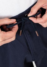 Marineblaue Hose mit einem Kordelzugbund, ausgestattet mit einem strukturierten, geflochtenen Bindeband und metallischen Spitzen. Nahaufnahme von Händen, die den Kordelzug anpassen.