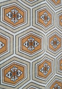 Hexagonaal stofpatroon met oranje en roomkleurige bloemen met zwarte omlijningen op een lichte achtergrond; geometrisch ontwerp met ingewikkelde details.