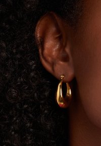 Gouden oorring met een curvilineair ontwerp, met een gladde, reflecterende oppervlakte. Bevestigd aan een kleine metalen sluiting.
