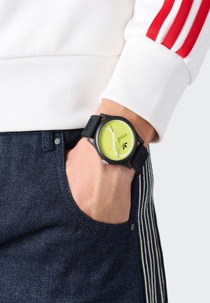 Juodas laikrodis su apvaliu ciferblatu, ryškiai žaliu skydeliu ir juodu silikono dirželiu, pasižymintis baltais rodyklėmis ir minimalistiniu dizainu.